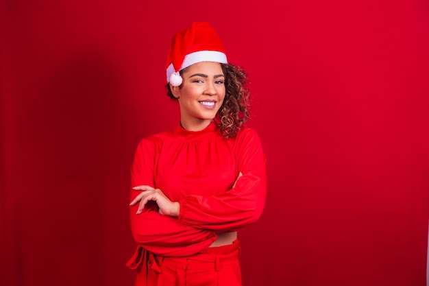 Afro-Frau trägt Weihnachtsmütze stehend vor rotem Hintergrund mit verschränkten Armen. isoliertes Porträt.