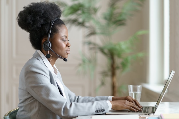 Afro-Geschäftsfrau im Blazer, der auf Schreibtisch sitzt, Fernarbeit am Laptop im Heimbüro, studiert