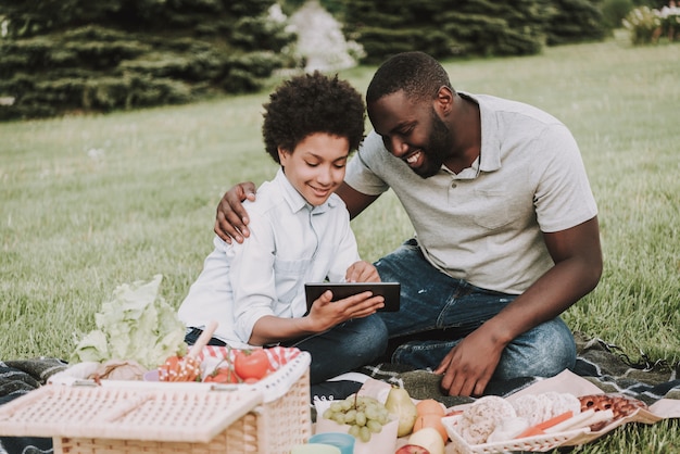 Afro hijo y padre mirando en tableta en Picnic