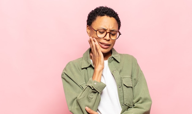 Afro joven mujer negra sosteniendo la mejilla y sufriendo dolor de muelas, sintiéndose enferma, miserable e infeliz, buscando un dentista