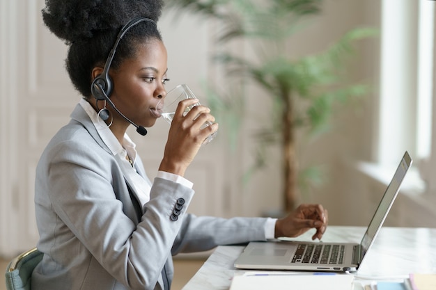 Afro-Mitarbeiterin, die Laptop-Bildschirm betrachtet, zu Hause arbeitend arbeitet, Wasser aus einem Glas trinkend.