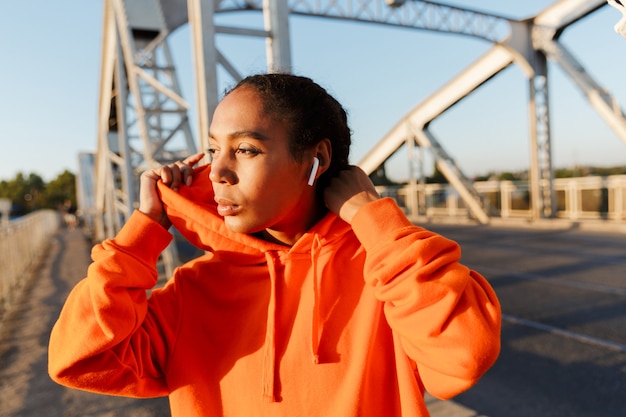 Afroamericana mujer satisfecha en ropa deportiva con earpods mientras se ejercita en el puente viejo