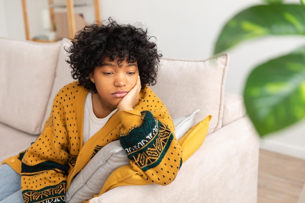 Afroamericana, triste, pensativa, pensativa, desmotivada, niña sentada en un sofá en casa interior. Una joven africana reflexiona, se ve cansada después de un largo día. La niña se siente deprimida, ofendida, solitaria, angustiada.