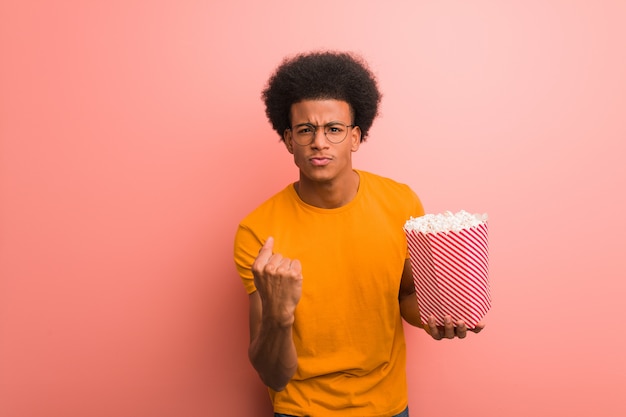 Afroamericano joven que sostiene un cubo de las palomitas de maíz que muestra el puño al frente, expresión enojada