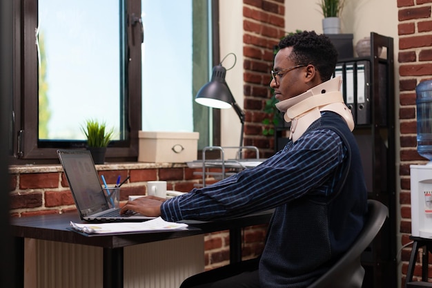 Afroamericano sentado en el escritorio en la oficina de inicio usando un collarín recuperándose después de un accidente trabajando en una computadora portátil. Empresario lesionado con collar cervical de regreso al trabajo después de un trauma físico.