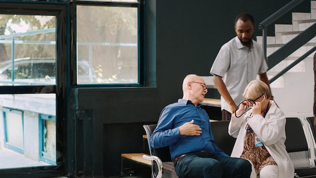 Afroamericanos ayudando a un anciano con problemas cardíacos a sentarse en el vestíbulo de recepción del hospital. Médico principal que consulta a un paciente mayor con estetoscopio, tratando de curar enfermedades cardiovasculares.