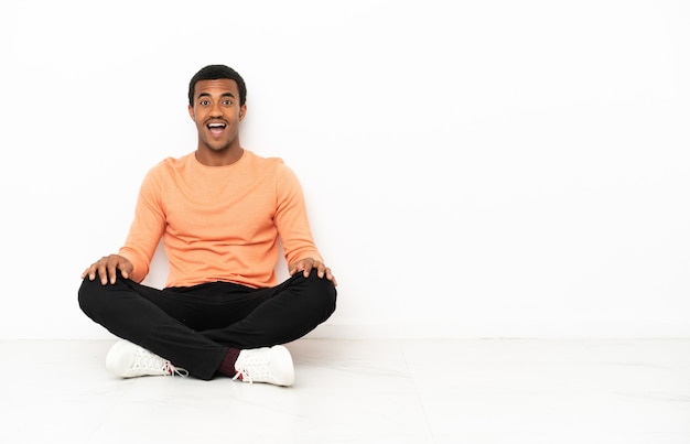Afroamerikaner, der auf dem Boden über isoliertem Hintergrund mit überraschendem Gesichtsausdruck sitzt