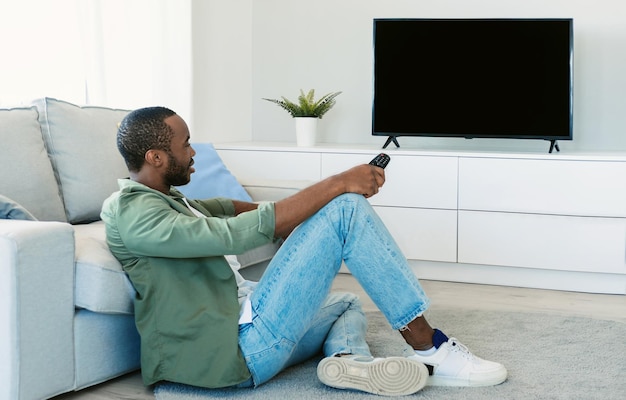Afroamerikaner, der fernsieht und die Fernbedienung auf ein leeres Bildschirmmodell zeigt