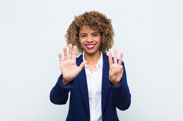 Afroamerikaner der jungen Frau, die freundlich lächelt und schaut, Nummer acht oder acht mit der Hand nach vorne zeigend, gegen flache Wand herunterzählend