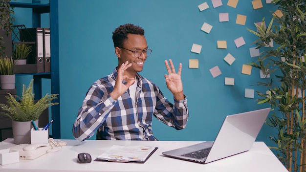 Afroamerikaner, der mit den Händen vor dem Laptop auf dem Schreibtisch ein Okay- und Genehmigungszeichen zeigt. Optimistischer Werbeerfolg für Erwachsene und ausgezeichnete Zufriedenheitsgeste.