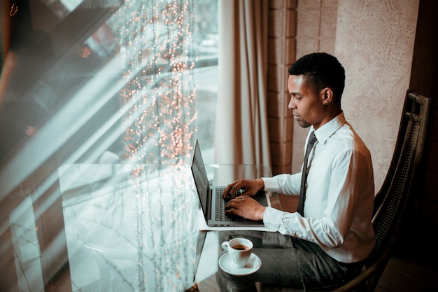 Afroamerikaner-Geschäftsmann, der auf Laptop arbeitet, gekleidet in weißem Hemd, Nahaufnahme, Innen-, Profilansicht, im Restaurant