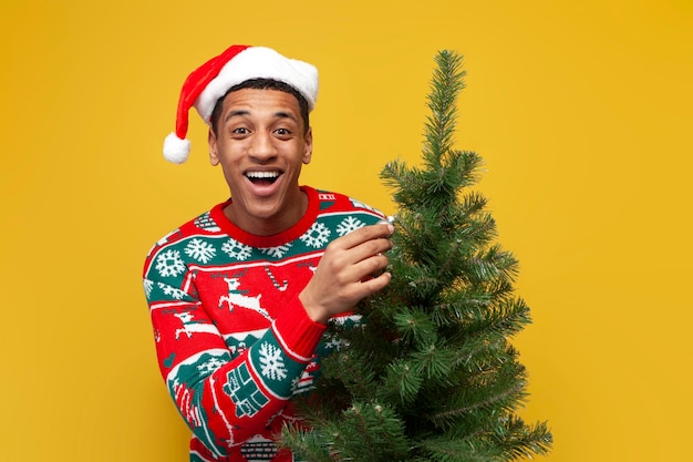 Afroamerikaner in Weihnachtskleidung und Weihnachtsmütze hält Weihnachtsbaum auf gelbem Hintergrund