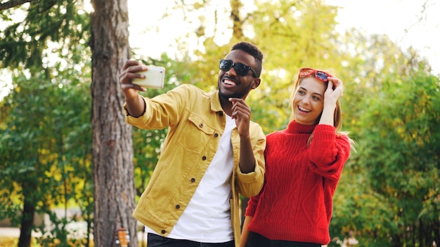 Afroamerikaner macht Selfie mit Freundin, die Smartphone hält und mit Sonnenbrille posiert, die zusammen im Park steht Freundschafts- und Technologiekonzept