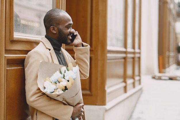 Afroamerikaner Mann in einer Stadt. Kerl, der Blumenstrauß hält. Männchen in einem braunen Mantel.