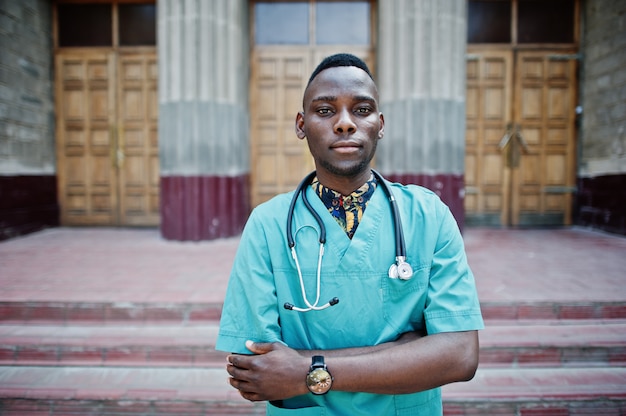 Afroamerikanerdoktormann am Laborkittel mit dem Stethoskop im Freien gegen Kliniktür.