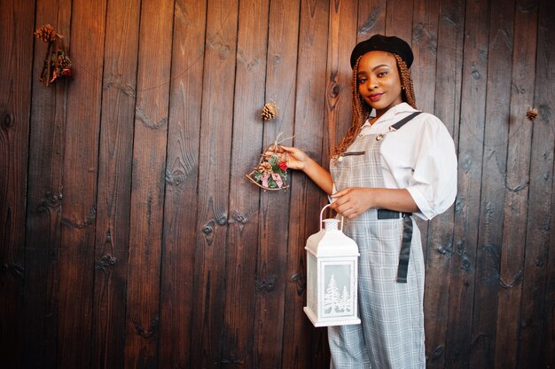 Afroamerikanerfrau in Overalls und Baskenmütze gegen Holzwand mit Laterne in der Hand.