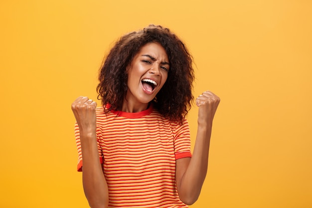 Afroamerikanerfrau mit Afro-Frisur, die erhobene Fäuste zusammenpresst, die über orange Wand zufrieden stehen