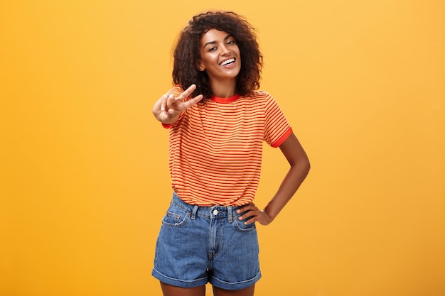 Afroamerikanerfrau mit lockiger Frisur, die Hand auf Hüfte hält und Kopf neigt, der freudig lächelnd Friedensgeste über orange Wand zeigt
