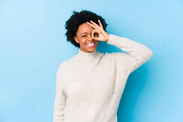 Afroamerikanerfrau mittleren Alters gegen eine aufgeregte blaue Wand, die ok Geste auf Auge hält.