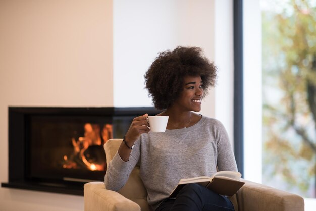 afroamerikanerfrau trinkt tasse kaffee lesebuch am kamin. Junges schwarzes Mädchen mit heißem Getränk, das sich beim Aufwärmen entspannt. Herbst zu Hause.