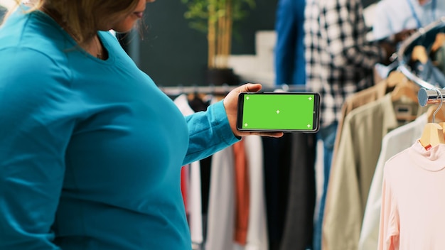 Afroamerikanerin analysiert Telefon mit Greenscreen-Vorlage und betrachtet isoliertes Display in moderner Boutique. Stilvoller Kunde kauft im Bekleidungsgeschäft ein trendiges Outfit. Modekonzept