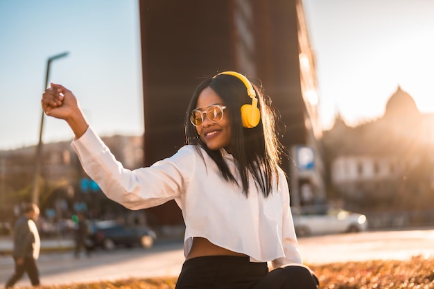 Afroamerikanerin, die bei sonnenuntergang in der stadtmusik online musik mit kopfhörern hört und lächelnd tanzt