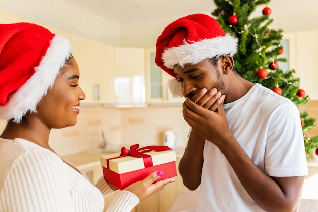 Afroamerikanerin, die ihrem Freund im Haus in der Nähe des Weihnachtsbaums eine Geschenkbox gibt