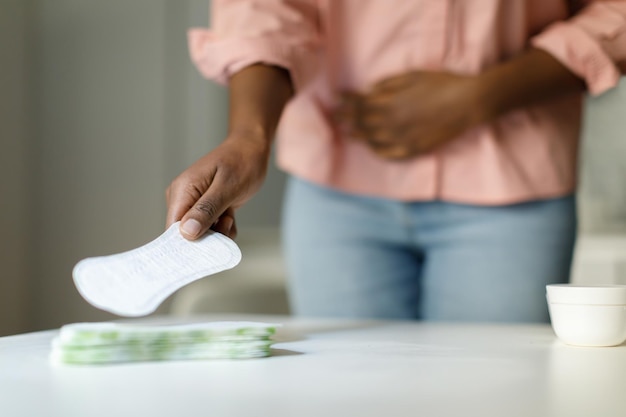 Afroamerikanerin, die während des Menstruationszyklus Schmerzen verspürt und täglich Damenbinden aus dem selektiven Fokus des Tisches nimmt