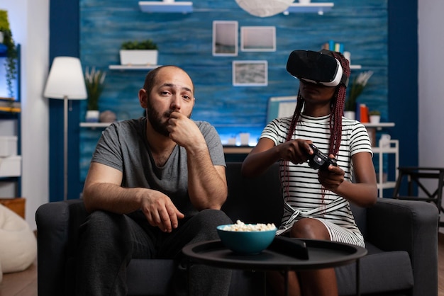 Afroamerikanerin genießt virtuelle Realität, während kaukasischer Mann Snacks beobachtet und isst. Junge Freundin spielt VR-Spiele mit High-Tech-Headset für simulierte Realität
