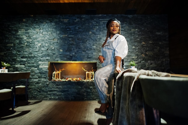 Afroamerikanerin im Overall und Baskenmütze im Zimmer mit Holztisch und Kamin