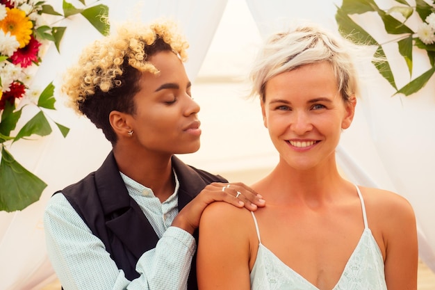 Afroamerikanerin in der Rolle des Bräutigams und der kurzhaarigen Braut im blauen Kleid in der Zeremonie unter dem Hochzeitsbogen am tropischen Strand