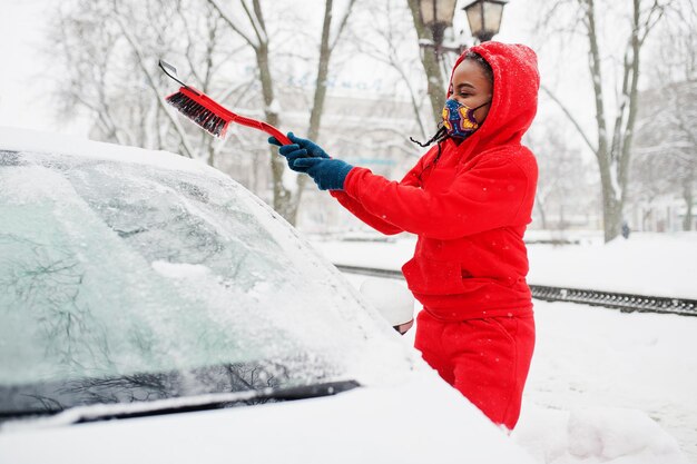 Afroamerikanerin in rotem Hoodie und Gesichtsmaske säubert das Auto am Wintertag vom Schnee.