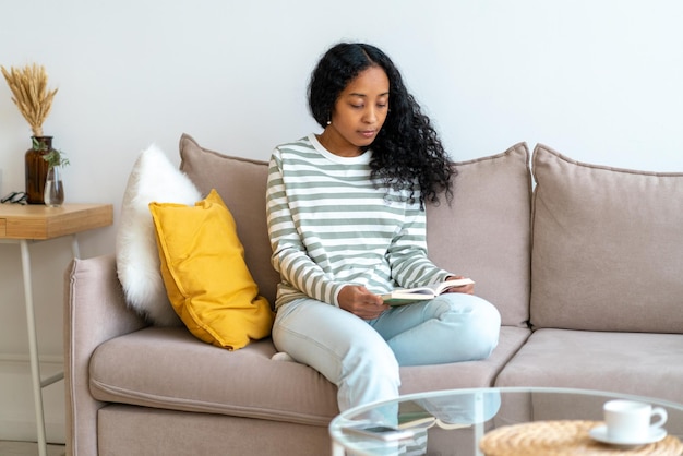 Foto afroamerikanerin liest ein buch, während sie im gemütlichen wohnzimmer auf dem sofa sitzt und zeit allein verbringt