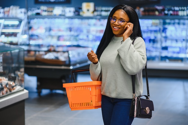 Afroamerikanerin mit Einkaufswagen im Supermarkt spricht mit dem Handy.