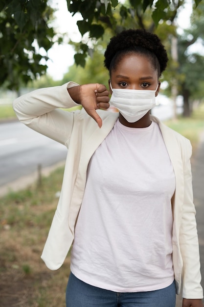 Foto afroamerikanerin mit medizinischer maske, die ein abneigungsschild mit daumen nach unten zeigt, das durch die straßen der stadt geht schlechter ausdruck