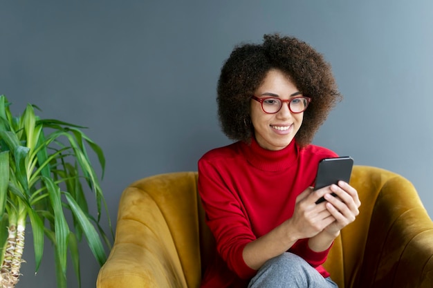 Afroamerikanerin mit Smartphone, die zu Hause auf einem Sessel sitzt und Videos ansieht