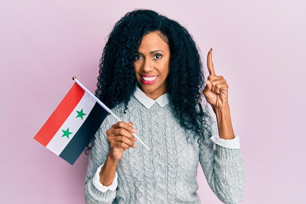 Afroamerikanerin mittleren Alters mit syrischer Flagge lächelnd mit einer Idee oder Frage, die mit dem Finger mit glücklichem Gesicht zeigt, Nummer eins