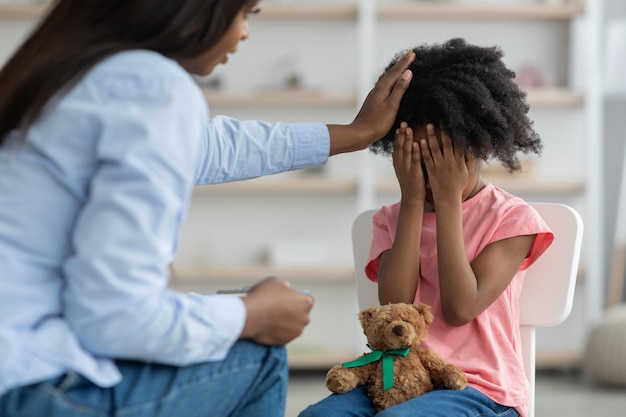 Afroamerikanerin tröstet emotionales, lockiges Mädchen im Vorschulklinik-Interieur