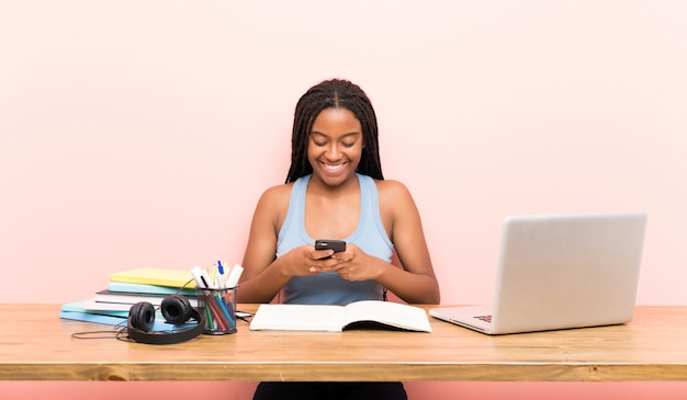 Afroamerikanerjugendlich-Studentenmädchen mit dem langen umsponnenen Haar an ihrem Arbeitsplatz, der eine Mitteilung mit dem Mobile sendet