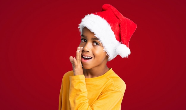 Afroamerikanerjunge mit Weihnachtshut etwas über rotem Hintergrund flüsternd