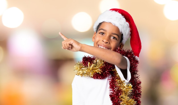 Afroamerikanerjunge mit Weihnachtshut zeigend auf die Seite, um ein Produkt über unscharfem Hintergrund darzustellen