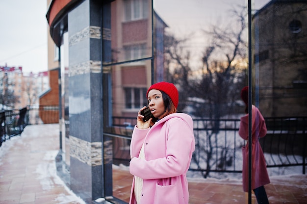 Afroamerikanermädchen im roten Hut und im rosa Mantel an der Straße der Stadt gegen das Errichten am Wintertag mit Handy.