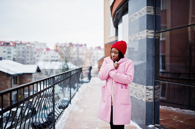 Afroamerikanermädchen im roten Hut und im rosa Mantel an der Straße der Stadt gegen das Errichten am Wintertag.