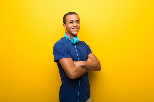 Afroamerikanermann mit blauem T-Shirt auf dem gelben Hintergrund, der die Arme gekreuzt hält