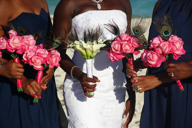 Afroamerikanische Braut auf weißem Kleid zusammen mit Brautjungfern, die bunte Blumensträuße am Hochzeitstag im Freien halten.