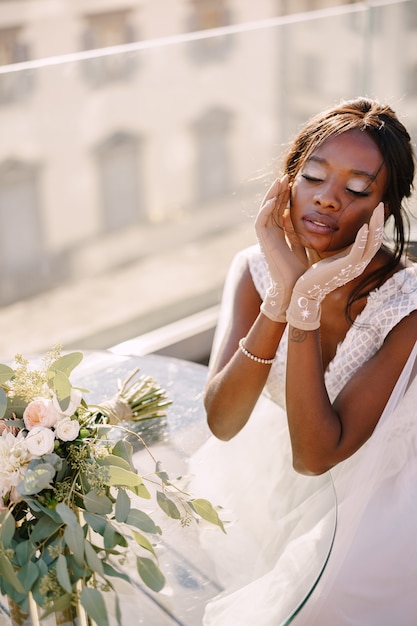 Afroamerikanische Braut sitzt am Tisch, berührt ihr Gesicht mit Handschuhen in den Händen, der Blumenstrauß liegt auf dem Tisch