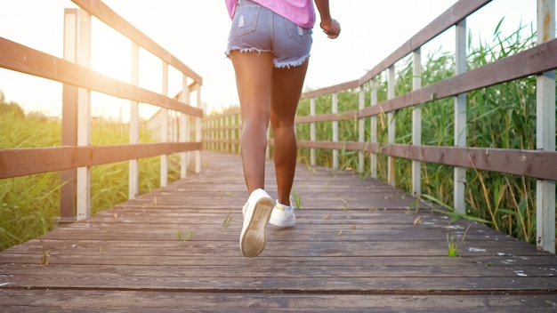 Afroamerikanische Dame mit langen, schlanken Beinen in blauen Shorts und heller Bluse läuft entlang einer Holzbrücke zwischen Feldern Rückansicht