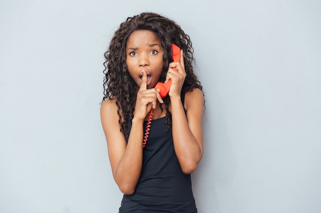 Afroamerikanische Frau, die auf Retro-Telefonröhre spricht und Finger über Lippen über grauer Wand zeigt