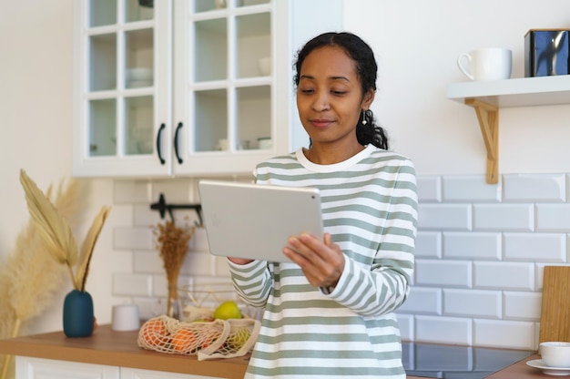 Afroamerikanische Frau, die ein digitales Gerät in der Küche verwendet, um ein Rezept zu suchen, bevor sie mit dem Kochen beginnt