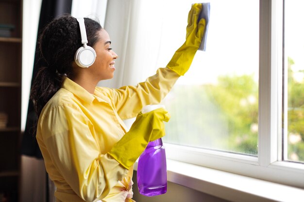 Afroamerikanische Frau, die Fensterglas mit Reinigungsmittelspray im Innenbereich putzt